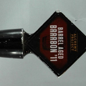 Puede incluir: Un tirador de cerveza negro con el texto "WIDMER BROTHERS" y "BARREL AGED BARBON '11". El tirador tiene un emblema cuadrado y una etiqueta en forma de diamante.