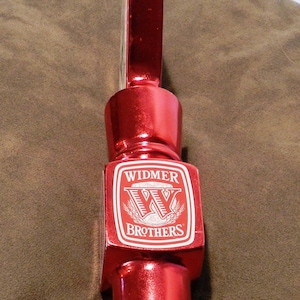 Puede incluir: Un brillante tirador de grifo de cerveza rojo con el logotipo "Widmer Brothers" en blanco. El tirador tiene una parte superior rectangular, una sección media cuadrada con el logotipo y una base cilíndrica acanalada. Diseñado para dispensar cerveza.
