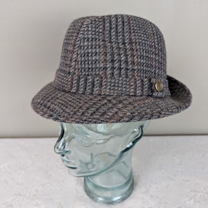 Può includere: Un cappello fedora a quadri grigi, marroni e rossi con un piccolo bottone dorato sul lato.