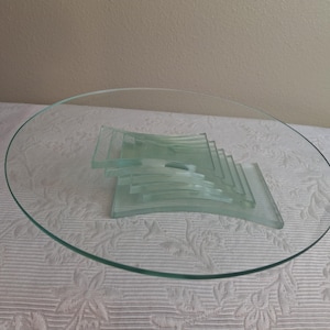 Puede incluir: Una bandeja de servicio de vidrio transparente con forma ovalada. La bandeja tiene una serie de piezas de vidrio curvadas apiladas en el centro. El vidrio tiene un ligero tinte verde. La bandeja está sobre una superficie blanca estampada.