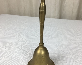 Pode incluir: Um sino de latão com uma longa alça e uma base alargada. O sino tem desenhos florais gravados ao redor da base. A alça é cilíndrica com anéis decorativos. O sino é de uma cor dourada quente.