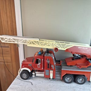 Op de afbeelding: Een rode speelgoed brandweerwagen met een gele en witte uitschuifbare ladder. De truck heeft zwarte banden, witte accenten en de tekst "DLK 23/12" aan de zijkant. Het staat op een wit oppervlak.