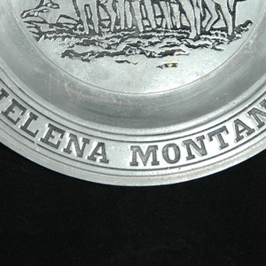 Puede incluir: Un plato de color plateado con las palabras "HELENA MONTANA" en relieve alrededor del borde. El plato presenta un relieve detallado de una escena hist&oacute;rica, probablemente representando un grupo de figuras. El plato es de metal.