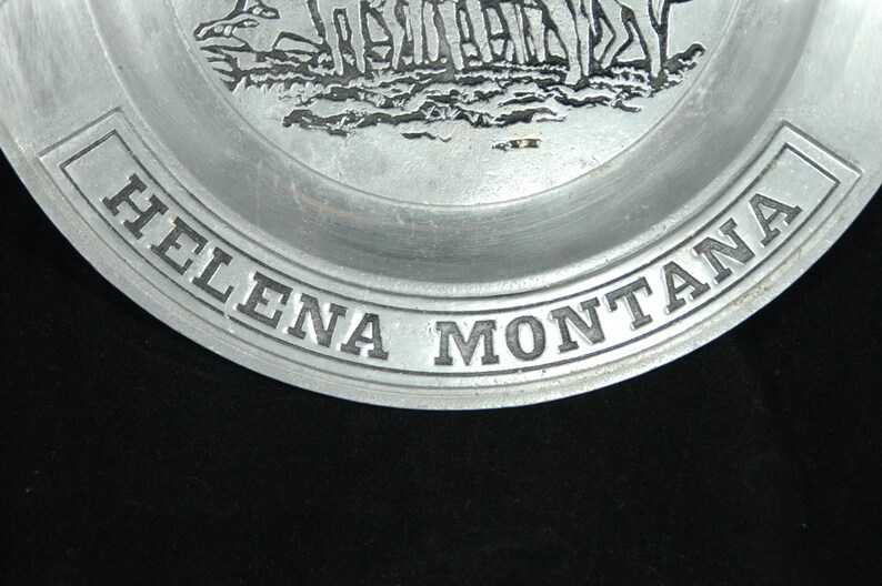 Puede incluir: Un plato de color plateado con las palabras "HELENA MONTANA" en relieve alrededor del borde. El plato presenta un relieve detallado de una escena hist&oacute;rica, probablemente representando un grupo de figuras. El plato es de metal.