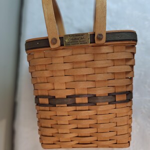 May include: A rectangular woven basket with two wooden handles. The basket is light brown with a dark brown band around the middle. A small metal plaque reads "Longaberger Collectors Club".