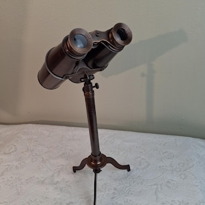 May include: Bronze-colored binoculars mounted on a tripod stand. The binoculars have a classic design with two lenses and a central focus mechanism. The tripod has three curved legs and a central support column.