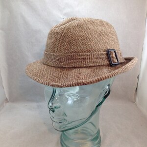 May include: A brown tweed hat with a herringbone pattern. The hat features a small brim and a band with a dark buckle. The hat is displayed on a clear glass head form.