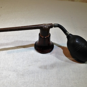 May include: A vintage brown glass perfume atomizer with a bulb. The atomizer has a long, straight nozzle and a bulb attached with a curved black tube. The bulb is black and has text on it.