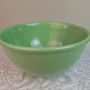 May include: A solid green ceramic mixing bowl. The bowl has a rounded shape with a slightly flared rim. The exterior is a smooth, glossy finish. The bowl is sitting on a white lace tablecloth.