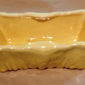 Puede incluir: Un plato rectangular de cerámica amarilla con un borde festoneado. El plato tiene un acabado brillante y un exterior ligeramente texturizado. El interior es liso y refleja la luz. Esta pieza decorativa podría usarse para objetos pequeños o como acento decorativo.