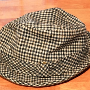 May include: A fedora-style hat with a black and tan checkered pattern. The hat has a structured crown and a brim that is slightly turned up. The fabric appears to be a woven material, and the hat is likely made for cooler weather.