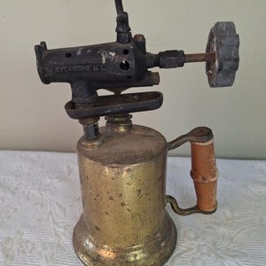 Op de afbeelding: Een antieke messing brander met een houten handvat. De brander heeft een zwarte metalen bovenkant met de tekst "SYCAMORE ILL". De handgreep is van hout en is bevestigd aan de messing tank. De brander is ontworpen voor solderen en andere verwarmingstoepassingen.