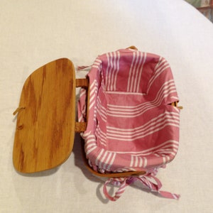 K&ouml;nnte beinhalten: Ge&ouml;ffneter Picknickkorb aus Holz mit rosa-wei&szlig; gestreiftem Stofffutter. Der Korb hat einen Klappdeckel und einen Griff. Der Stoff ist mit einem rosa Band gebunden. Der Korb ist leer.