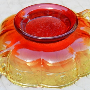 May include: An orange and yellow glass bowl with a circular, raised center. The bowl has a scalloped edge and decorative floral patterns. The glass has a glossy finish, with a gradient of colors from yellow to orange.