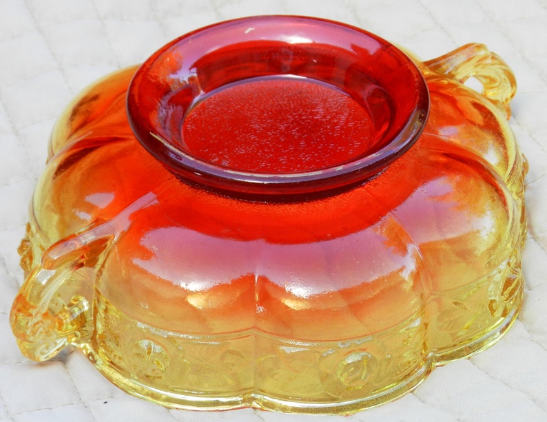 May include: An orange and yellow glass bowl with a circular, raised center. The bowl has a scalloped edge and decorative floral patterns. The glass has a glossy finish, with a gradient of colors from yellow to orange.