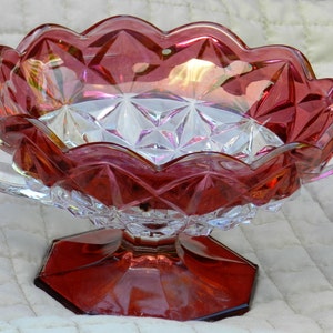 May include: A ruby-colored, scalloped-edge glass bowl with two clear handles. The bowl has a faceted design and sits on a hexagonal base. The interior of the bowl has a diamond pattern. A decorative piece for serving or display.