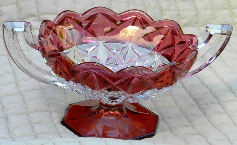 May include: A ruby-colored, scalloped-edge glass bowl with two clear handles. The bowl has a faceted design and sits on a hexagonal base. The interior of the bowl has a diamond pattern. A decorative piece for serving or display.