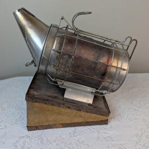 May include: A vintage metal bee smoker with a wire cage and a spout. The smoker is sitting on a wooden base.