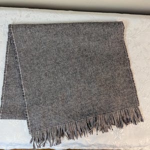 May include: A gray and white woven scarf with fringe on the ends. The scarf is folded in half and laid flat on a white surface.