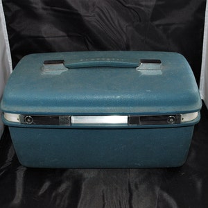 May include: A teal blue hard-sided travel case with a silver handle and silver accents. The case has a rectangular shape and a smooth surface.