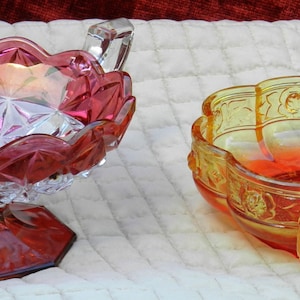 May include: Two decorative glass bowls. One is ruby red with a scalloped rim and a pedestal base. The other is amber with a floral design and handles. Both are on a white quilted surface.