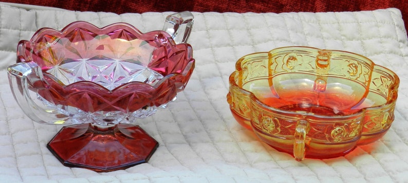 May include: Two decorative glass bowls. One is ruby red with a scalloped rim and a pedestal base. The other is amber with a floral design and handles. Both are on a white quilted surface.