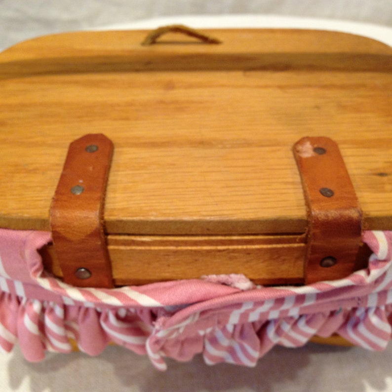 K&ouml;nnte beinhalten: Ein Vintage-Picknickkorb aus hellem Holz. Der Korb hat einen Klappdeckel, Lederriemen und ein rosa-wei&szlig; gestreiftes Stofffutter mit ger&uuml;schtem Rand. Ein kleiner Griff ist am Deckel befestigt.