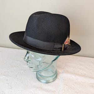 May include: A black fedora hat with a black band and a small feather accent. The hat is sitting on a mannequin head.