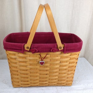 May include: A woven wicker basket with two wooden handles and a red fabric liner. The basket has a red heart charm hanging from it.