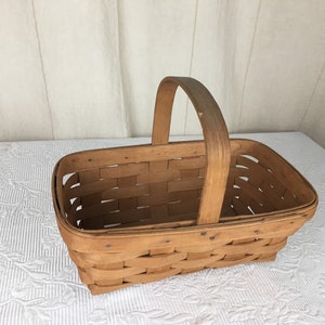 May include: A rectangular, light brown woven basket with a wooden handle. The basket is made of woven strips of wood and has a sturdy handle for carrying. The basket is likely used for storage or display.
