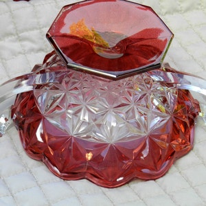 May include: A vintage red and clear glass serving dish with a geometric pattern. The dish has a scalloped edge and two clear arched handles. The base is a deep red, while the upper portion is clear.