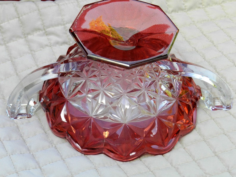 May include: A vintage red and clear glass serving dish with a geometric pattern. The dish has a scalloped edge and two clear arched handles. The base is a deep red, while the upper portion is clear.