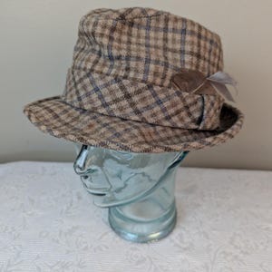 May include: A brown and blue plaid fedora hat with a small feather accent. The hat is on a mannequin head.