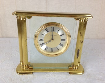 May include: A gold-toned clock with Roman numeral markings on a white face. The clock is encased in a glass and brass frame with decorative columns.