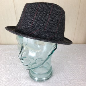 May include: A grey fedora hat with a herringbone pattern and thin red stripes. The hat has a short brim and a structured crown. The hat is displayed on a clear glass head form.