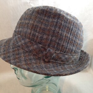 May include: A tweed cloche hat in shades of brown, gray, and blue. The hat features a structured brim and a decorative band around the crown. The fabric has a textured appearance.