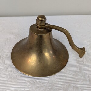May include: A vintage brass bell with a curved handle and a rounded base. The bell has a warm, golden hue and sits on a white patterned surface. The handle is attached to the top with a nut.