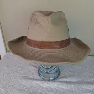 May include: A tan-colored hat with a wide brim and a brown leather band. The hat is made of a durable fabric and has a classic, outdoorsy style. The hat is sitting on a glass object.