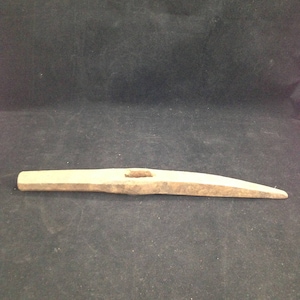 May include: A weathered, wooden tool with a rectangular body and a tapered, pointed end. The tool has a central, rectangular hole and appears to be made of a light-colored wood. The background is a dark, textured surface.