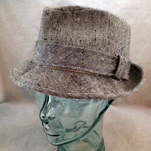 May include: A gray tweed fedora hat with a dark band and a small bow detail. The hat is displayed on a clear glass head form. The hat has a classic, structured shape with a slightly curved brim.