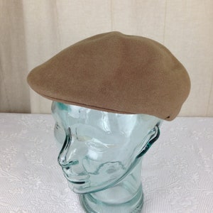 May include: A tan-coloured flat cap with a rounded crown and a small, stiff brim. The hat is made of a soft, felt-like material. The cap is displayed on a clear glass head form.