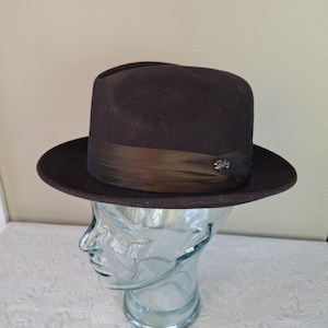 Pode incluir: Um chapéu fedora castanho escuro com aba larga e faixa de fita castanha escura. O chapéu é exibido em uma cabeça de vidro transparente. Um pequeno emblema prateado está preso à fita.