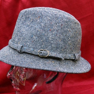 Puede incluir: Un sombrero fedora de tweed azul con ala pequeña y una banda decorativa con hebilla. El sombrero tiene una forma clásica y está hecho de una tela texturizada con motas de color.