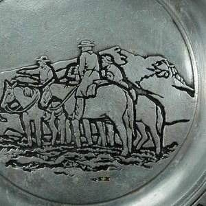 Puede incluir: Un plato plateado con un relieve detallado de tres figuras a caballo, atravesando un paisaje monta&ntilde;oso. El borde del plato presenta un dise&ntilde;o geom&eacute;trico. El relieve es de estilo vintage.