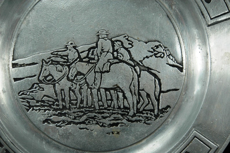 Puede incluir: Un plato plateado con un relieve detallado de tres figuras a caballo, atravesando un paisaje monta&ntilde;oso. El borde del plato presenta un dise&ntilde;o geom&eacute;trico. El relieve es de estilo vintage.