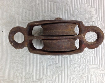 May include: A rusty metal double pulley with two round wheels and two loops on either side.