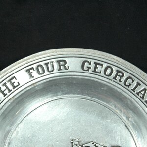 Puede incluir: Un plato de color plateado con las palabras "THE FOUR GEORGIANS" en relieve en el borde. El plato tiene un dise&ntilde;o simple y cl&aacute;sico, con un fondo oscuro.