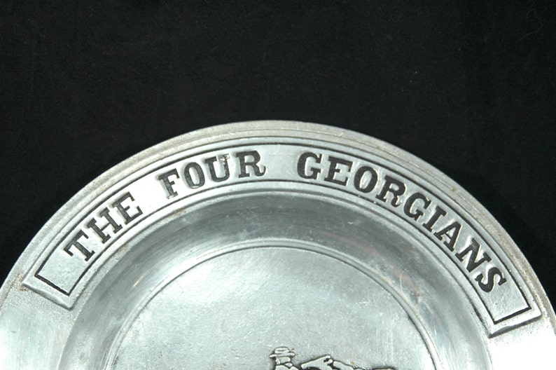 Puede incluir: Un plato de color plateado con las palabras "THE FOUR GEORGIANS" en relieve en el borde. El plato tiene un dise&ntilde;o simple y cl&aacute;sico, con un fondo oscuro.