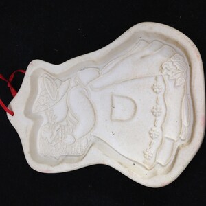 Può includere: Stampo per biscotti in ceramica bianco sporco con un rilievo dettagliato di una persona con cappello e vestito, che tiene un cestino. Lo stampo ha un nastro rosso per appenderlo. Il design include elementi floreali e la lettera "D".