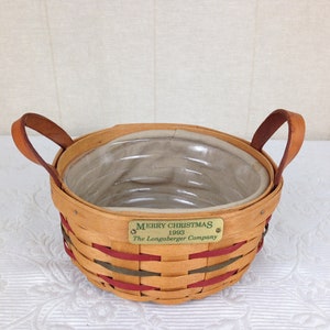 May include: A brown wicker basket with red and green accents and leather handles. The basket has a white fabric liner and a brass tag that reads "Merry Christmas 1993 The Longaberger Company."